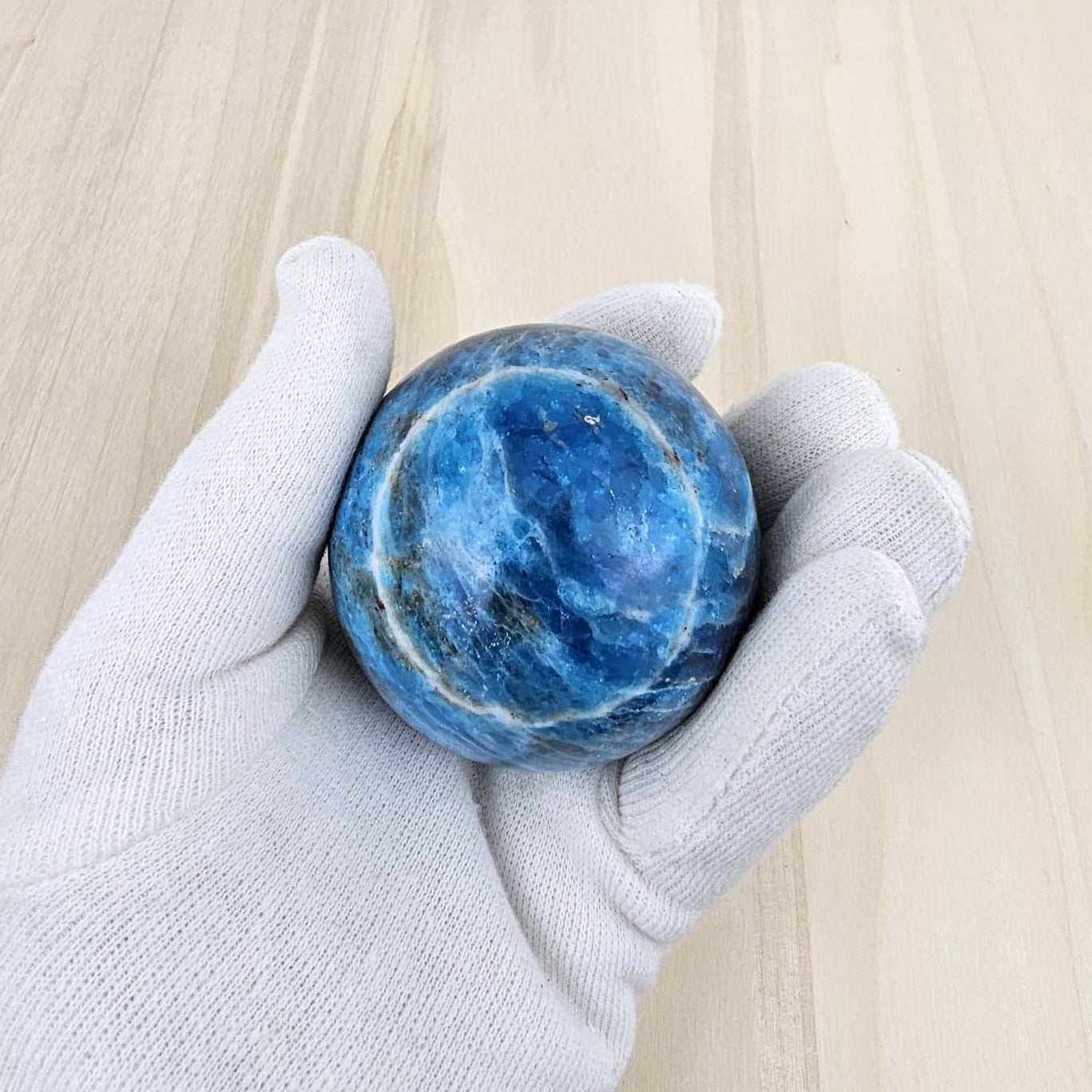 Blue apatite sphere held in a gloved hand on a light wooden surface