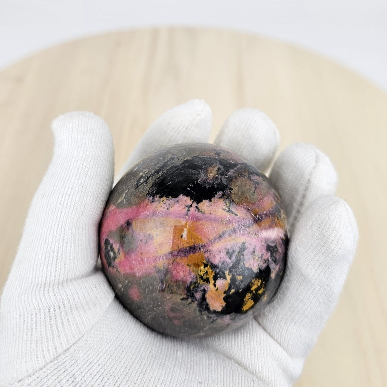 rhodonite crystal sphere held by a gloved hand on a beige surface with a white background