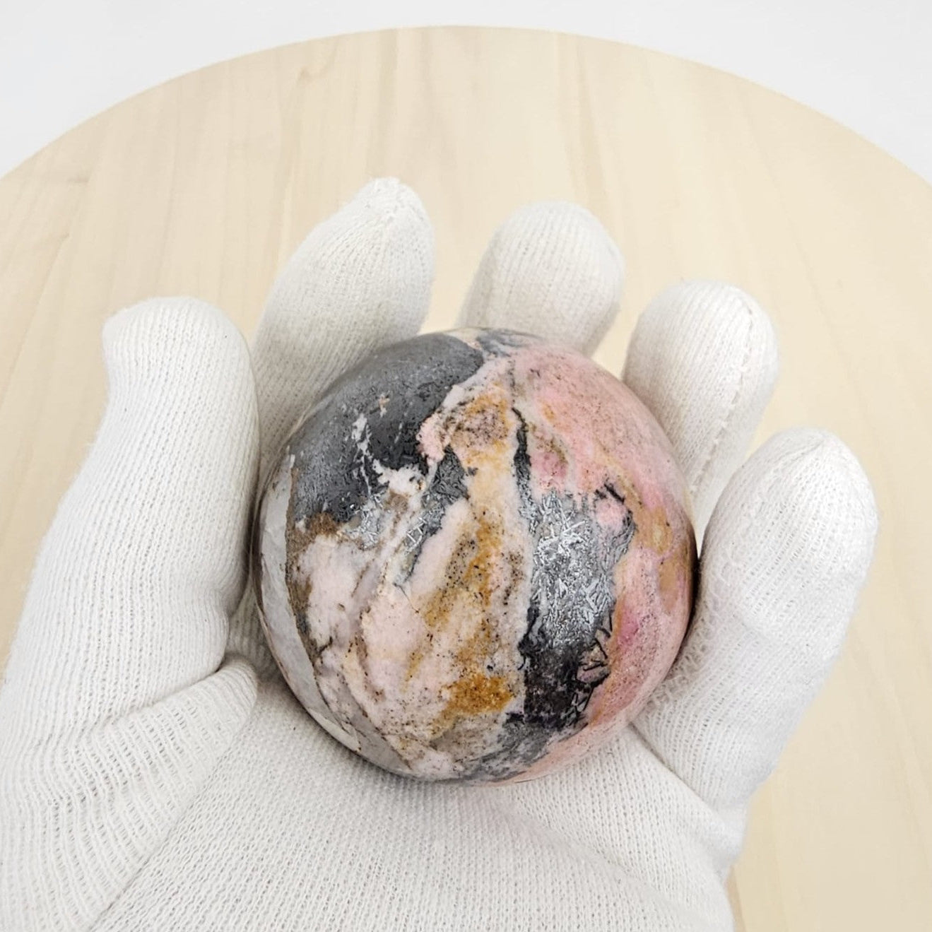 rhodonite crystal sphere held in a white glove on a light wooden surface